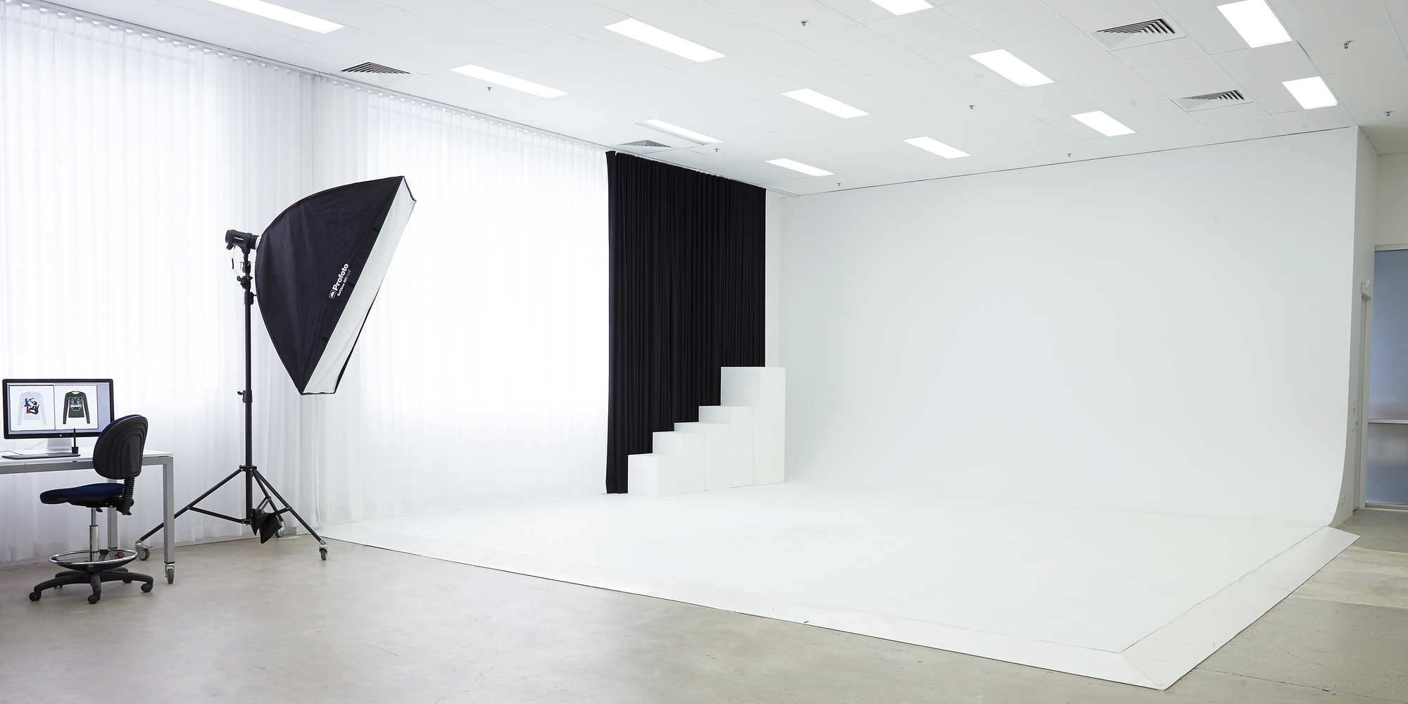 Photo studio setup with softbox lighting, computer monitor, white backdrop, and black curtain for professional photography.