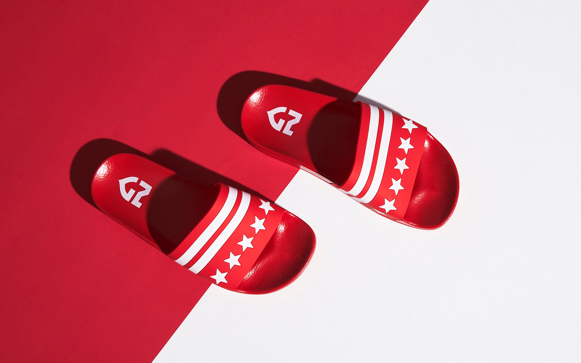 Red slide sandals with white stripes and stars on a red and white background, top-down view, product shot, casual footwear.