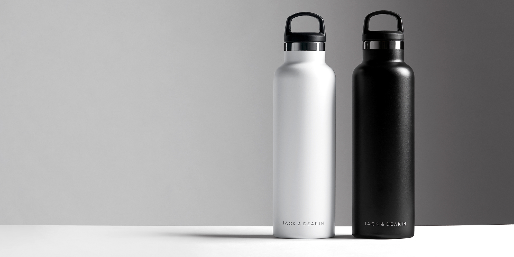 Two sleek Jack & Deakin water bottles, one white, one black, stand side-by-side on a white surface against a blended gray backdrop.