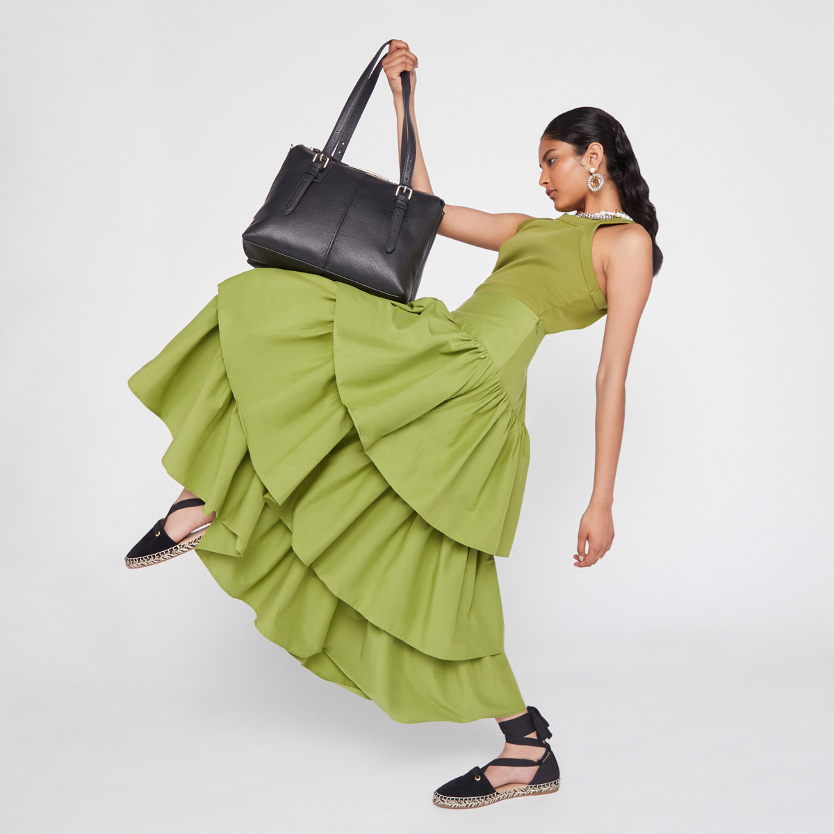 A woman in a green dress and black shoes poses in a studio holding a black leather handbag by the handle, with a white background.
