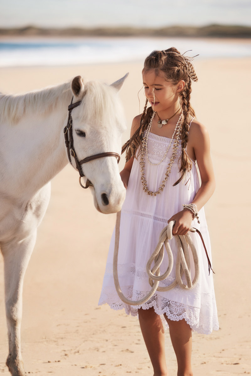 A young girl in a white dress stands on a beach next to a white pony, holding its rope lead on a sunny day near the ocean.
