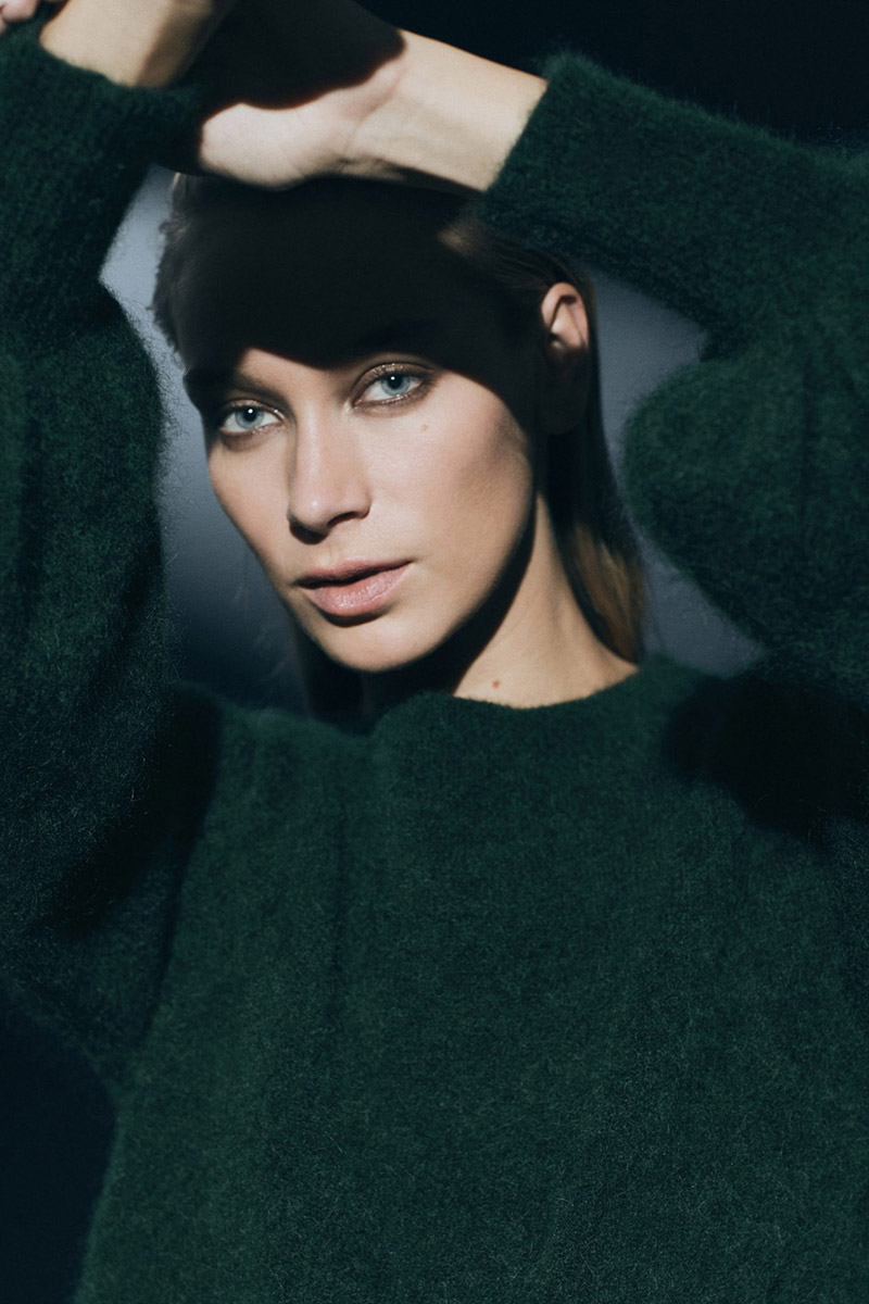 Portrait of a young woman with fair skin and blue eyes, wearing a dark green sweater, posing with her hands above her head.