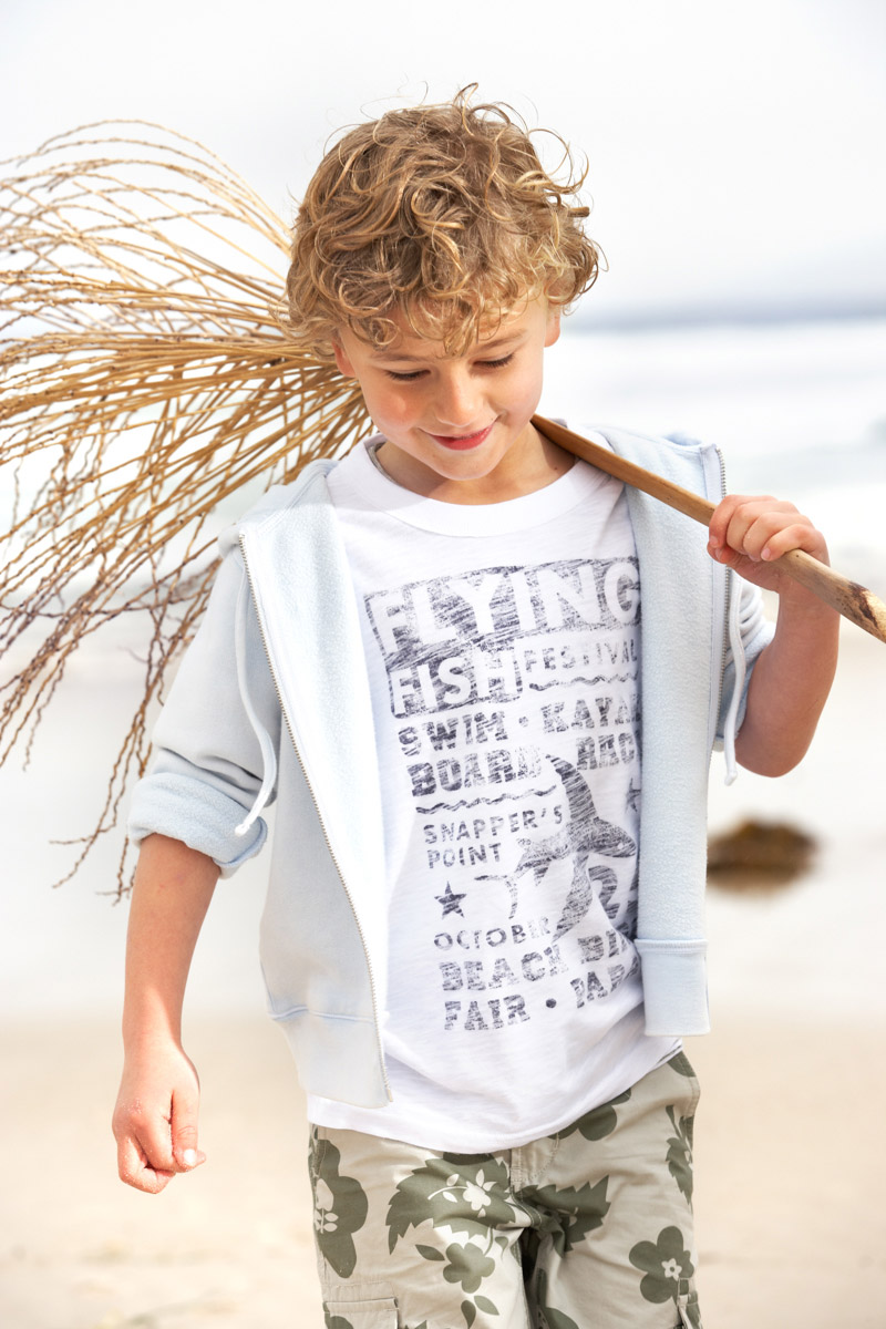 A young boy with curly blonde hair walks on a beach carrying a stick with foliage, wearing a t-shirt and floral shorts.