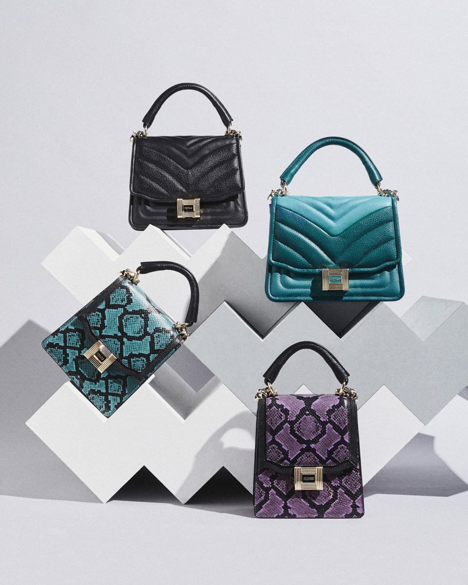 Four fashionable handbags in black, blue, teal, and purple snake print arranged on a white geometric platform against a gradient gray backdrop.
