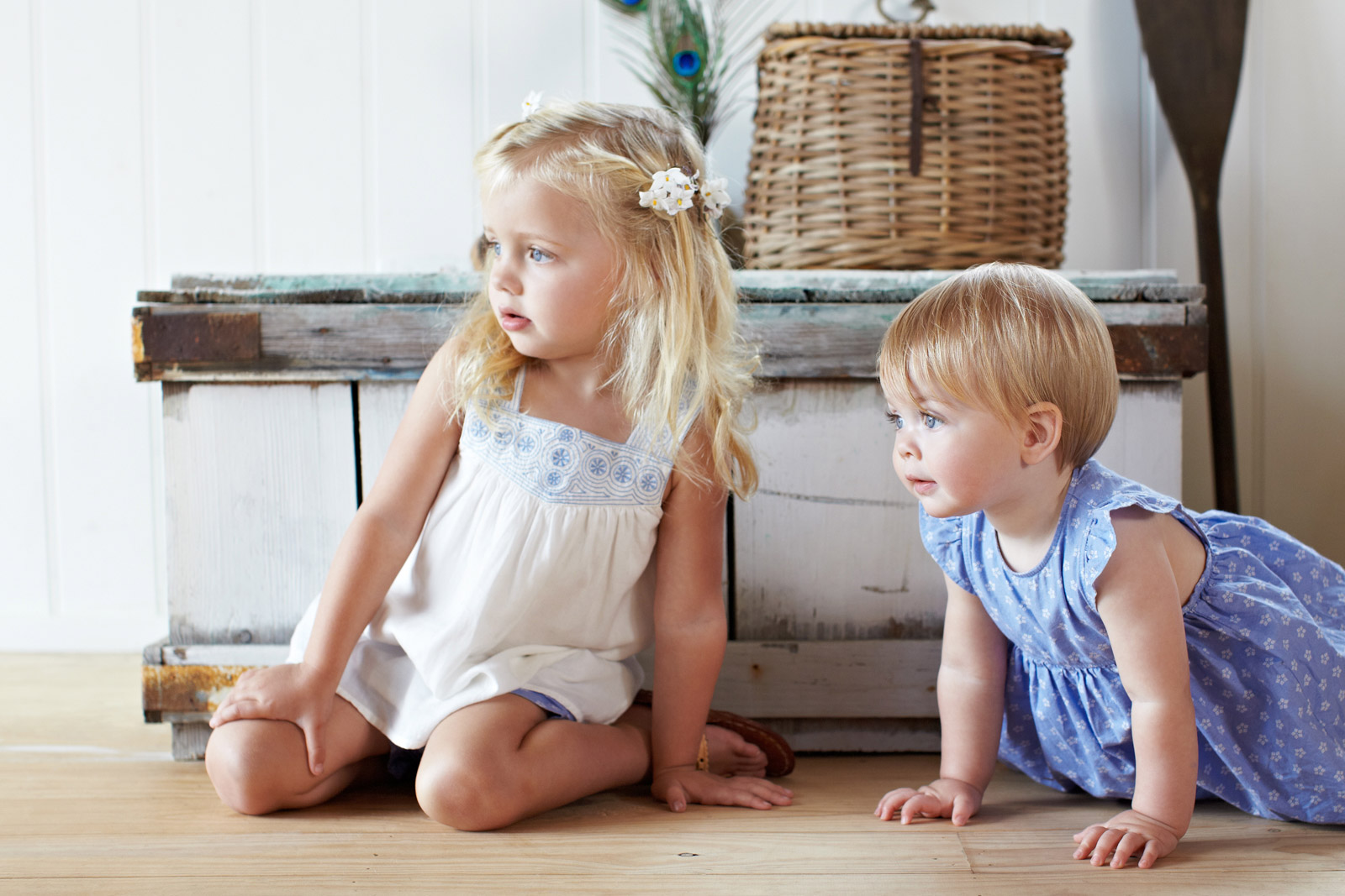 Two young children, one sitting and one crawling, in a bright, indoor setting with natural light and a rustic wooden backdrop and floor.