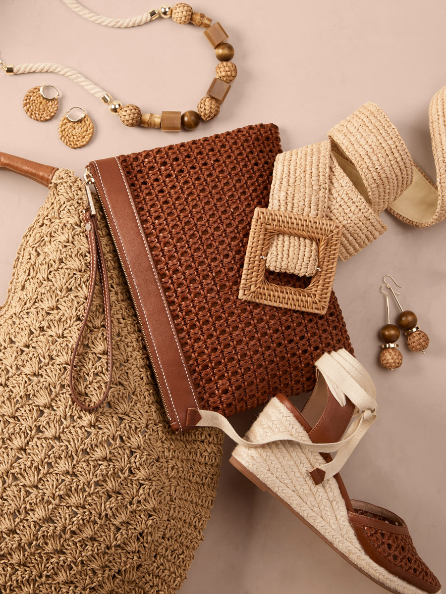 Still life of summer fashion accessories including a woven straw tote bag, clutch, earrings, necklace, belt, and espadrille platform shoes.