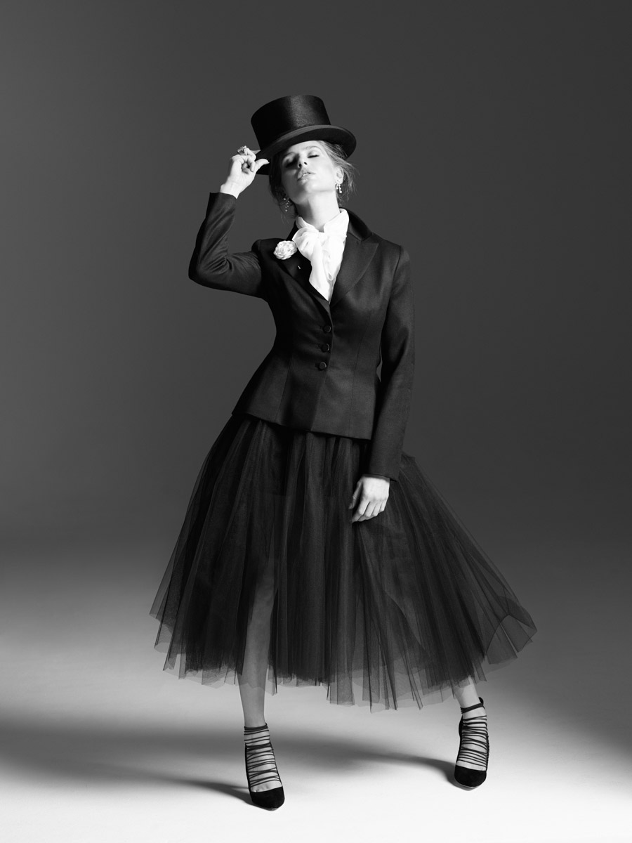 Black and white portrait of a woman in a suit and tutu, tipping a top hat while posing in a photo studio with soft lighting.
