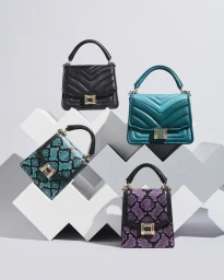 Four handbags in black, teal, turquoise snakeskin, and purple snakeskin displayed on geometric white pedestals.