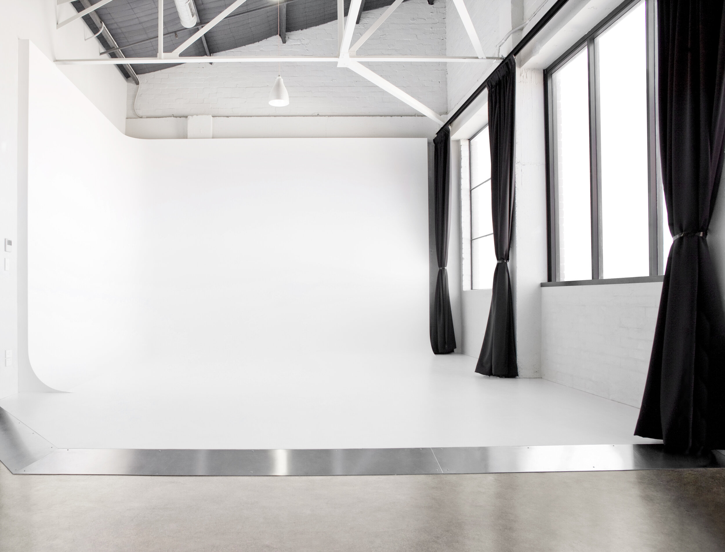 Empty photo studio with white cyclorama wall, natural light from windows with black curtains, and a concrete floor.