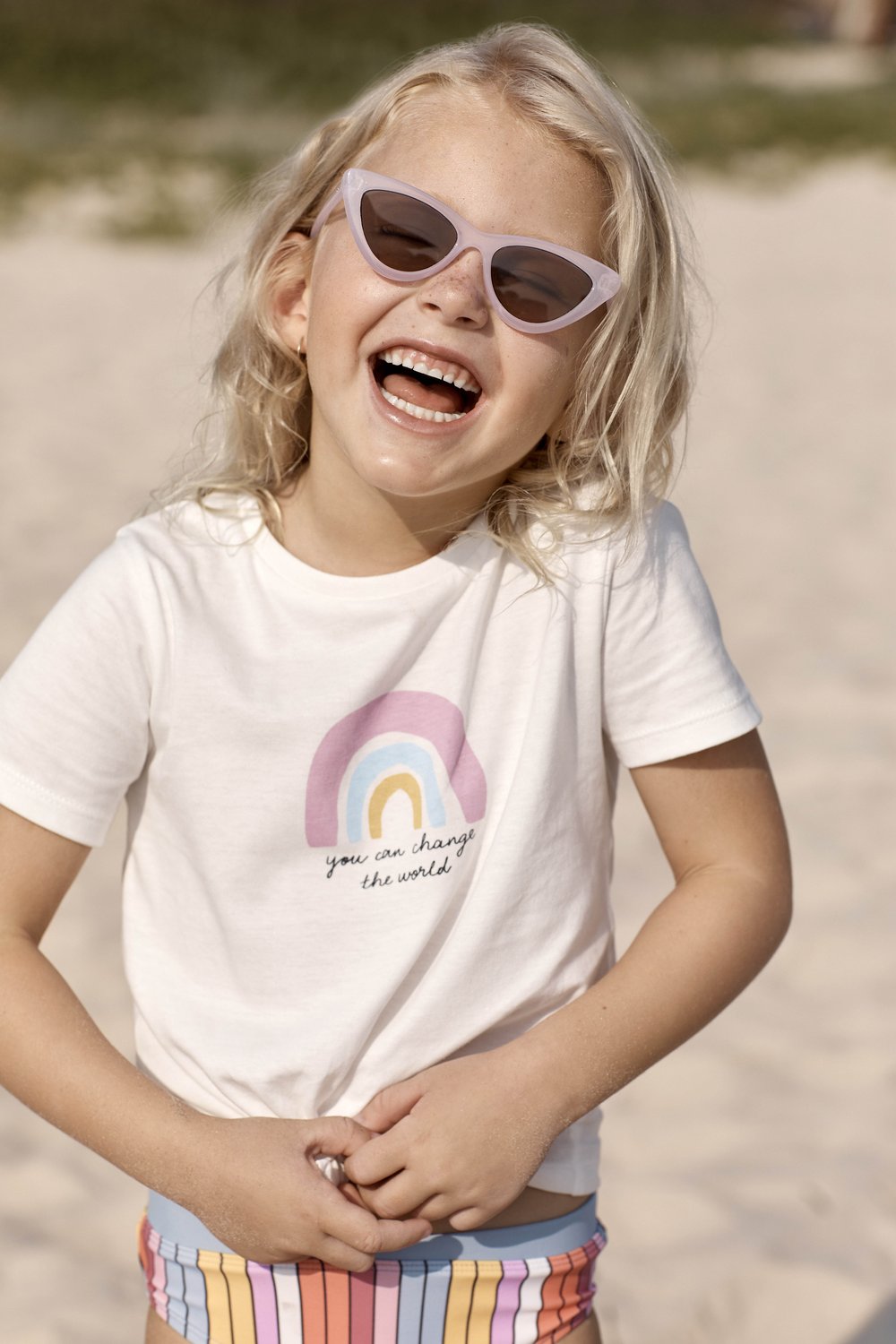 Happy young girl with blond hair wearing sunglasses and a t-shirt with a rainbow graphic on a sandy beach setting.