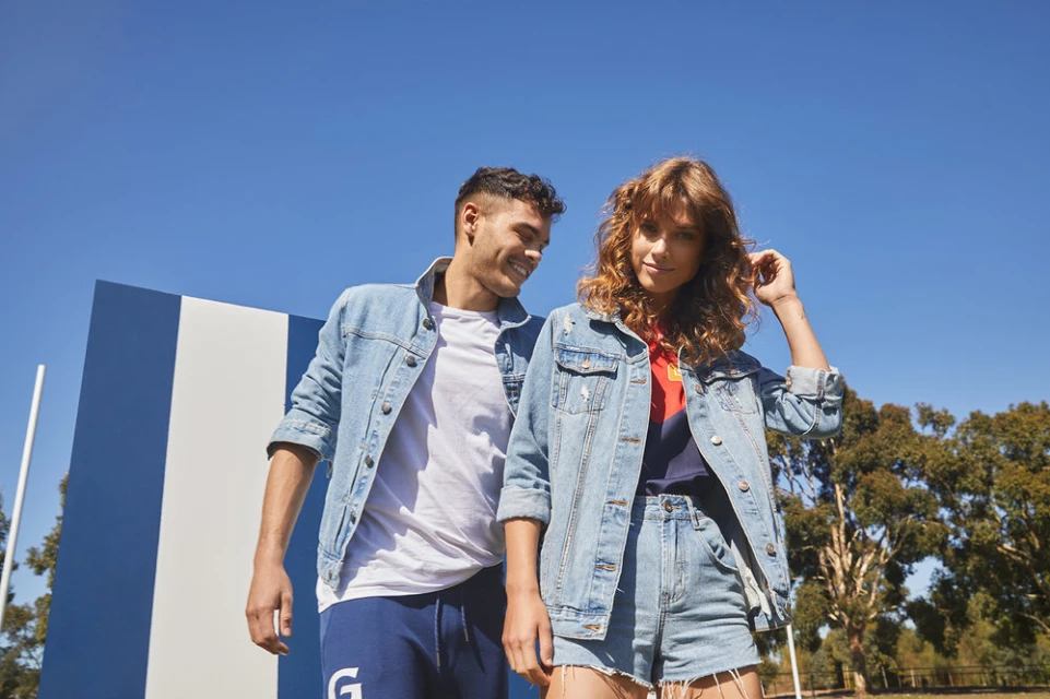 Outdoor shot of a stylish couple wearing denim jackets and shorts under a clear blue sky, with an urban background and greenery.