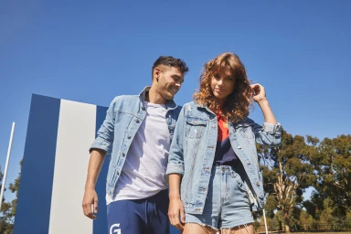 A man and woman wearing matching denim jackets standing outdoors under a bright blue sky with trees and a panel structure in the background.