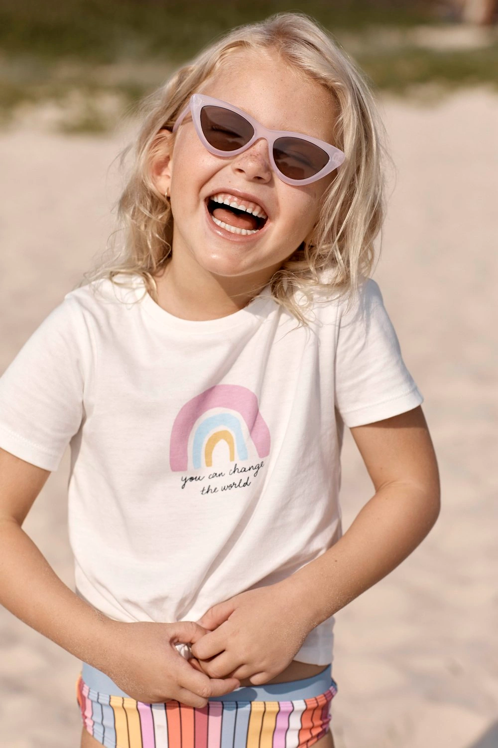 Young blond child laughing outdoors wearing pastel pink cat-eye sunglasses and a white t-shirt with a rainbow graphic.