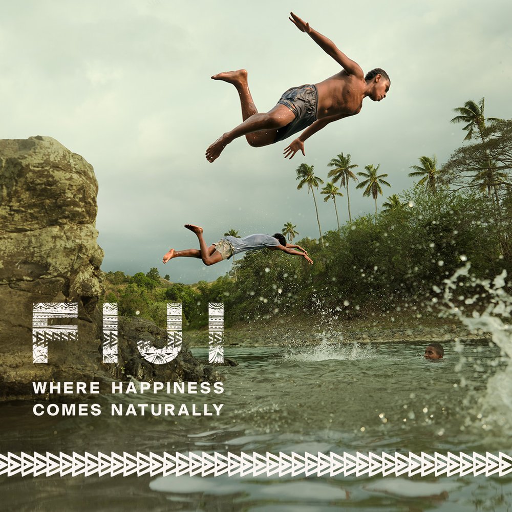 Two people dive into tropical water near a rocky outcrop, with a background of palm trees and a cloudy sky in an island setting.