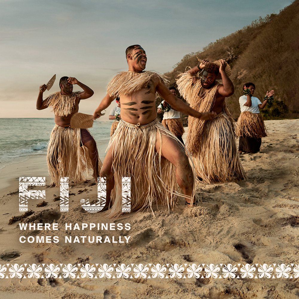 Men in traditional Fijian dress dance on a sandy beach with lush green mountain backdrop, under a bright sky at dusk.