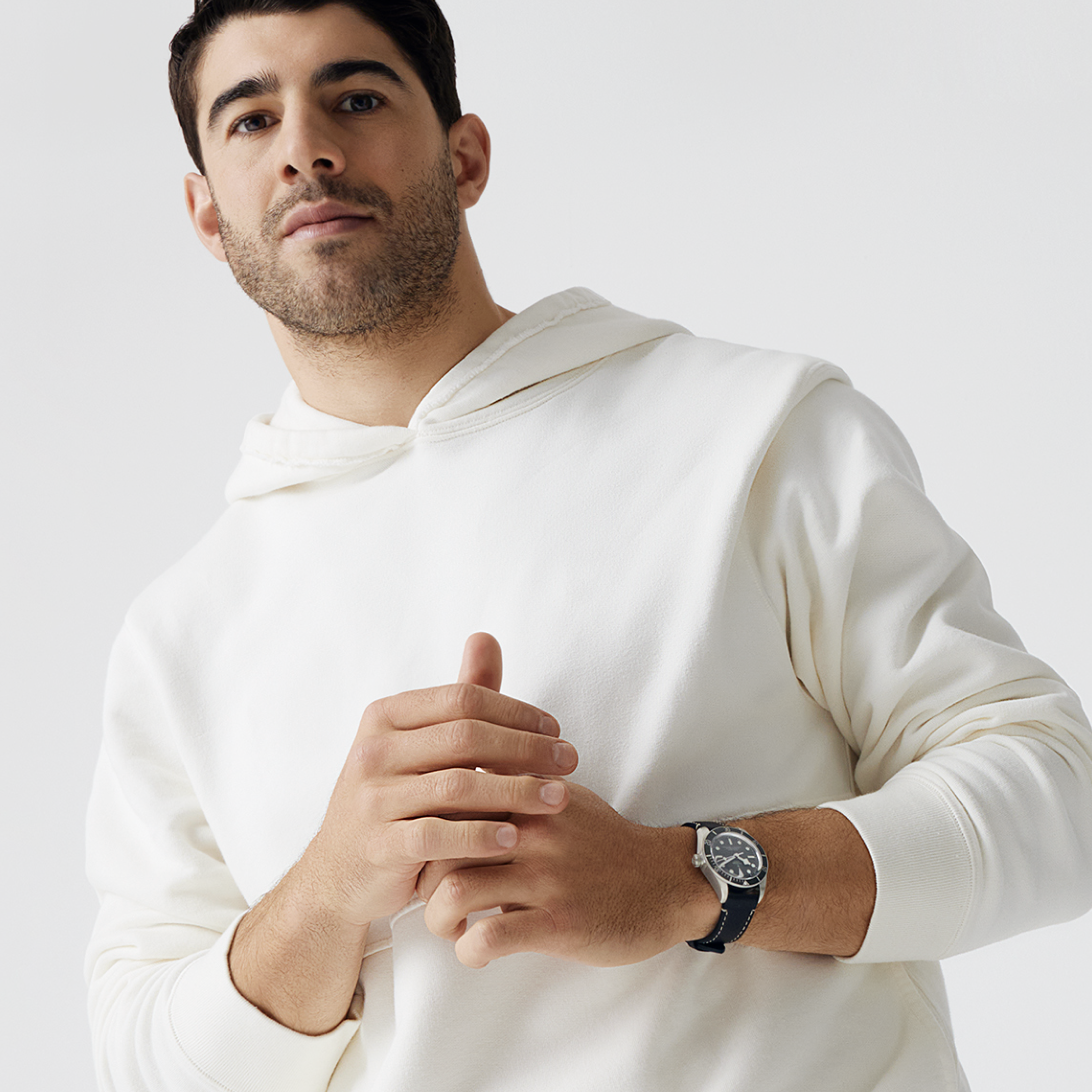 Portrait of an adult male wearing a white hoodie and wristwatch with black strap, standing indoors against a neutral studio backdrop.