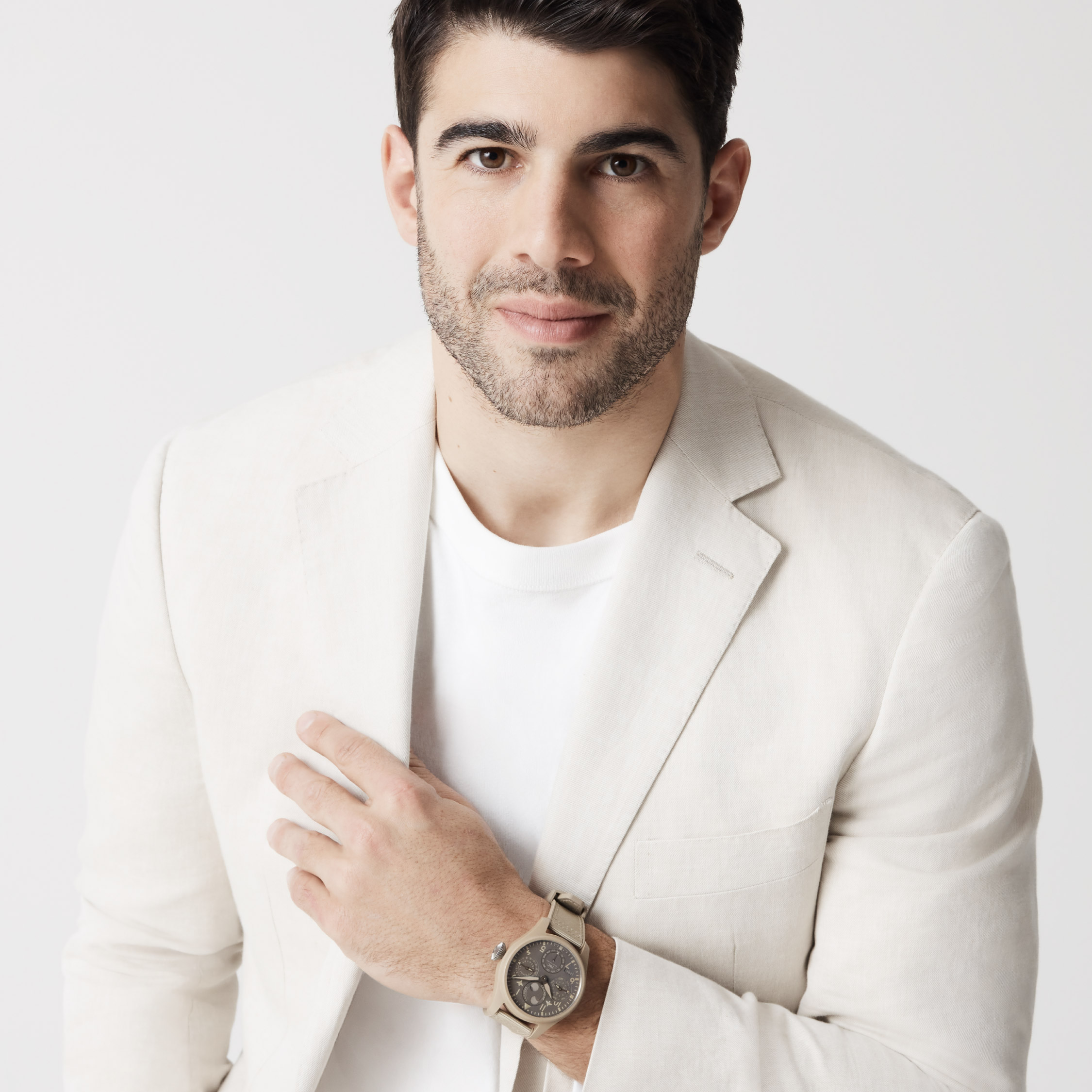 Portrait of a man in a cream suit and white t-shirt, wearing a watch, posing against a light backdrop in a photo studio setting.