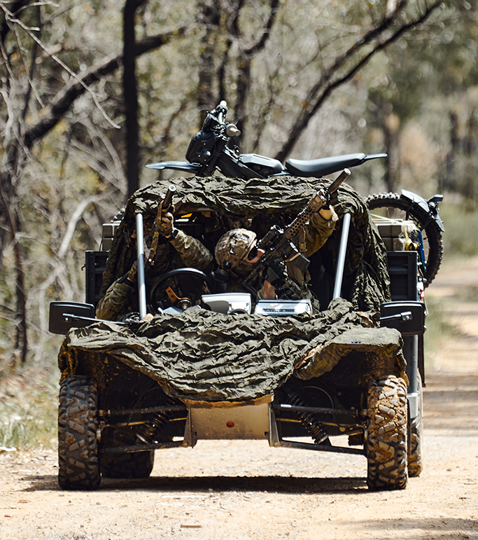 Three people in camouflage tactical gear ride in an all-terrain vehicle on a dirt road surrounded by a forest of trees.