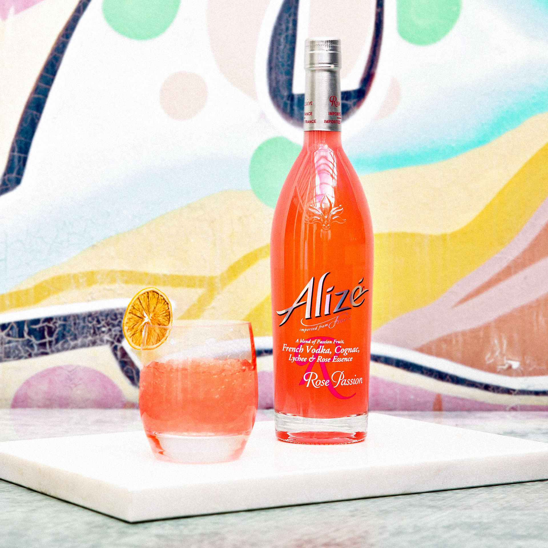 Alize Rose Passion bottle next to a cocktail in a glass with an orange slice garnish, set against an abstract painted backdrop.