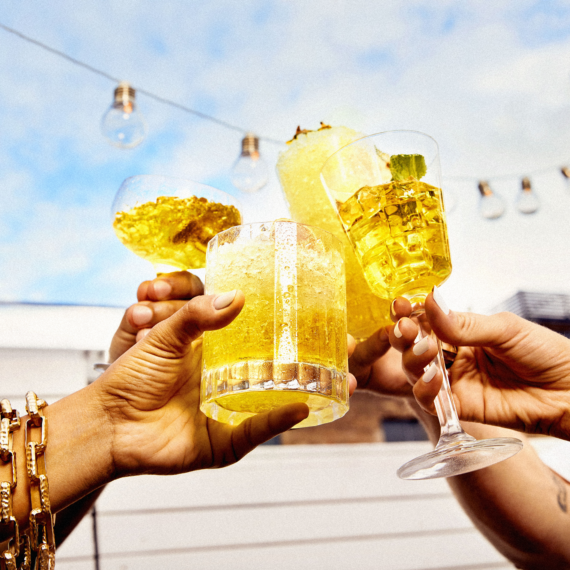 A group of people toasting with yellow cocktails in various glasses, under string lights, creating a celebratory and vibrant outdoor scene.