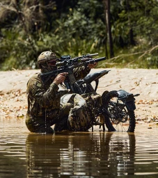 Soldier in camouflage uniform crouching in water next to a dirt bike while aiming a rifle