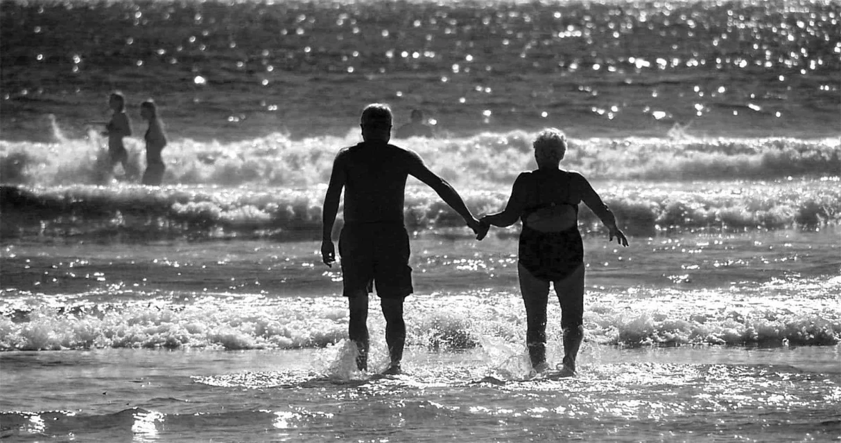 Elderly couple walking hand-in-hand on a beach with gentle waves in black and white, creating a timeless and serene seaside scene.