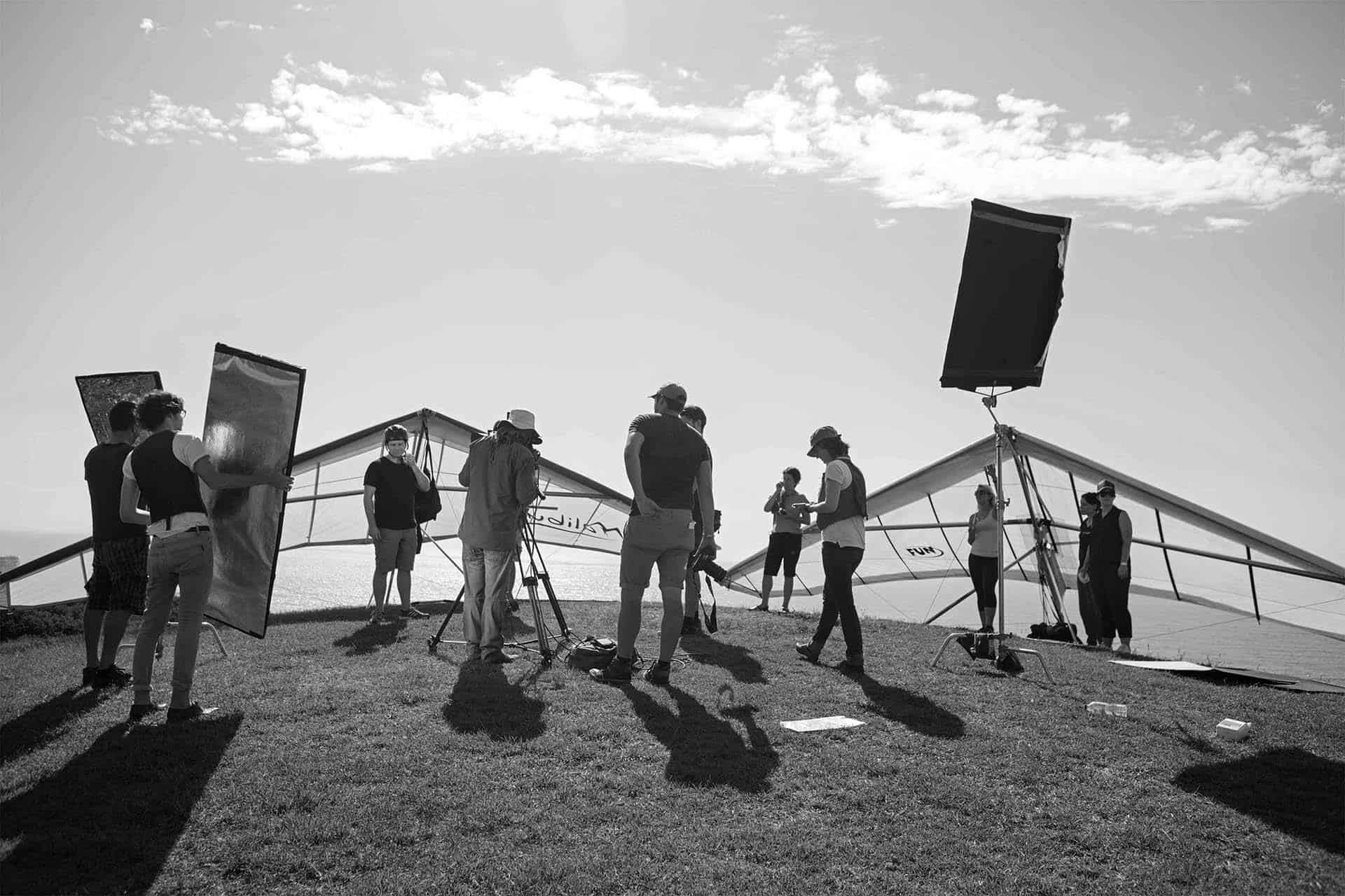 Black and white image of a film crew with lighting equipment, cameras, and hang gliders on a grassy hilltop against a cloudy sky.