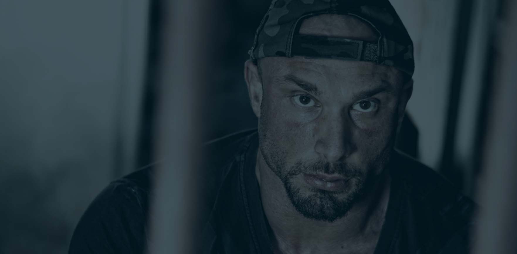 Close-up portrait of a muscular adult male with a camouflage cap, medium beard, and serious expression in a dimly lit, indoor setting.