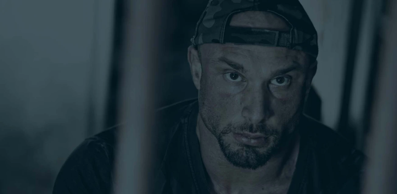 Close-up portrait of a muscular adult male with a camouflage cap, medium beard, and serious expression in a dimly lit, indoor setting.