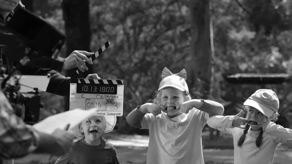 Black and white photo of three children making faces on film set with crew holding slate board in front of trees on a sunny day.
