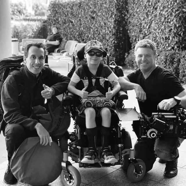 Black and white shot of a child in a wheelchair between two men, all giving thumbs-up signs, in an airport-like setting near bushes.