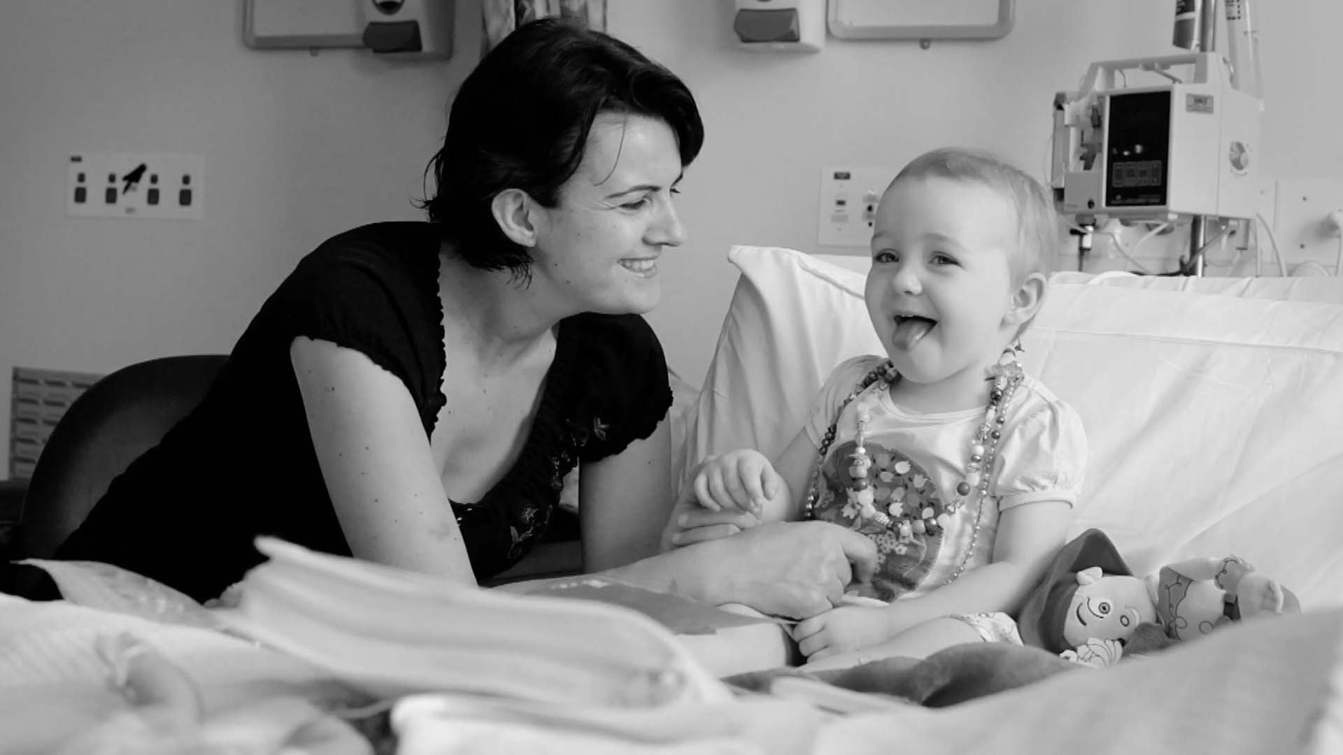 Black and white photograph of a woman smiling at a toddler sitting on a hospital bed surrounded by toys, creating a heartwarming scene.