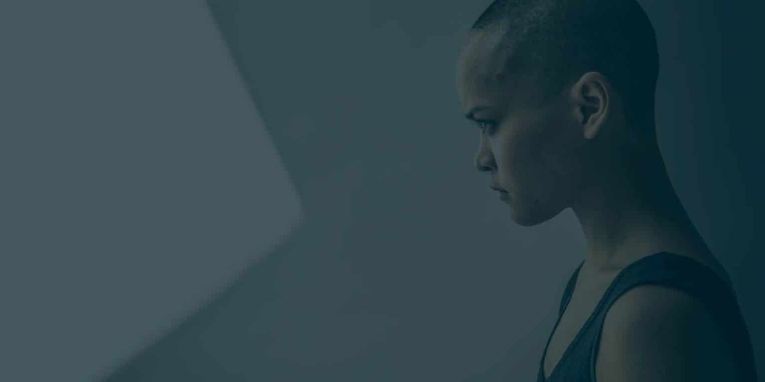 Striking portrait of a woman with a shaved head in profile, against a muted grey background, emphasizing her determined expression.