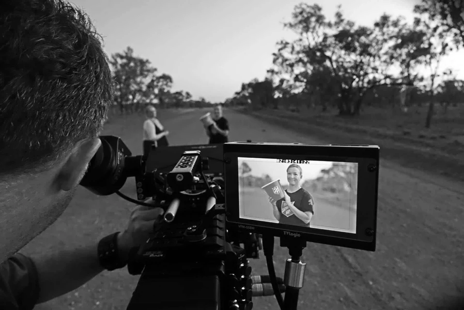 Film crew recording scene in rural location with a camera operator, two adult actors, outdoor setting, and natural lighting.
