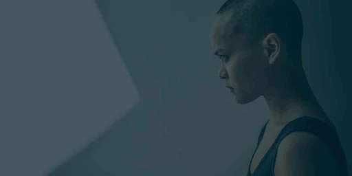 Profile portrait of a person with a shaved head wearing a dark tank top in profile view against a dimly lit blue-toned background.