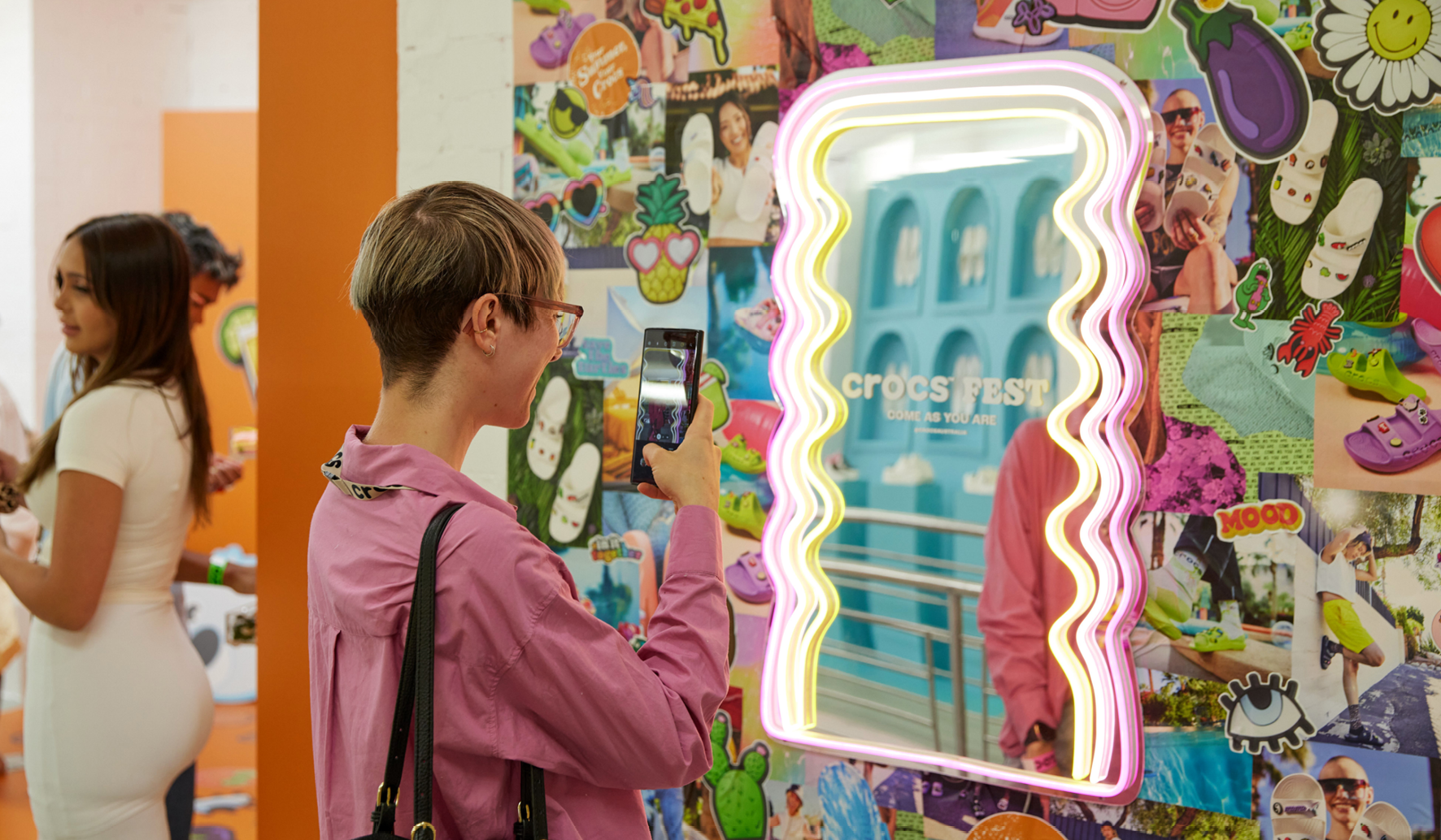 Woman photographs Crocs Fest display featuring shoes and cartoon imagery, framed by neon lights, in an indoor setting with other people.