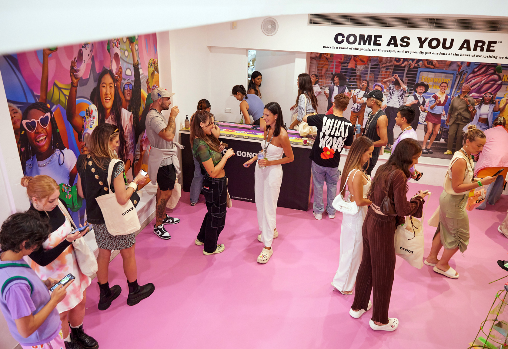 A group of people are gathered inside a pink Crocs store with an illustration on the wall, looking at their phones and mingling.