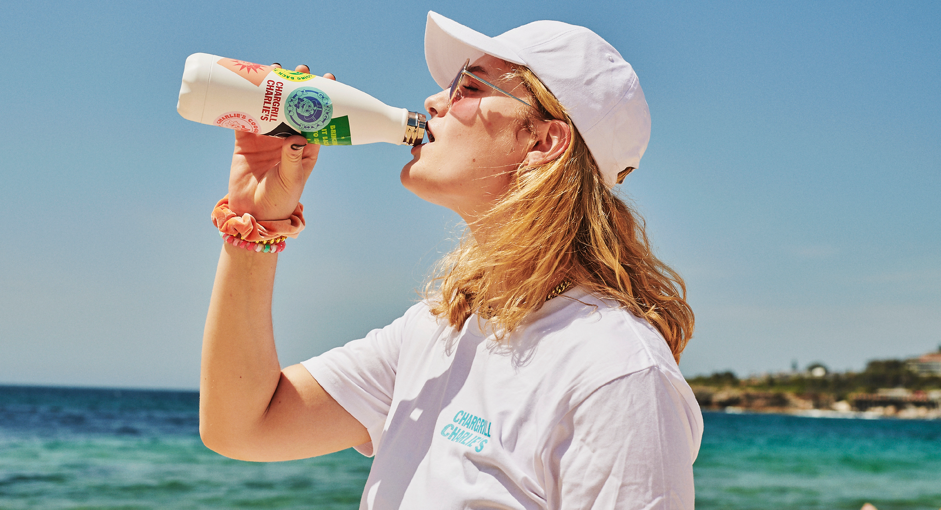 Young woman in sunglasses and baseball cap drinking from a bottle at a beach on a sunny day with bright blue sky and ocean view.