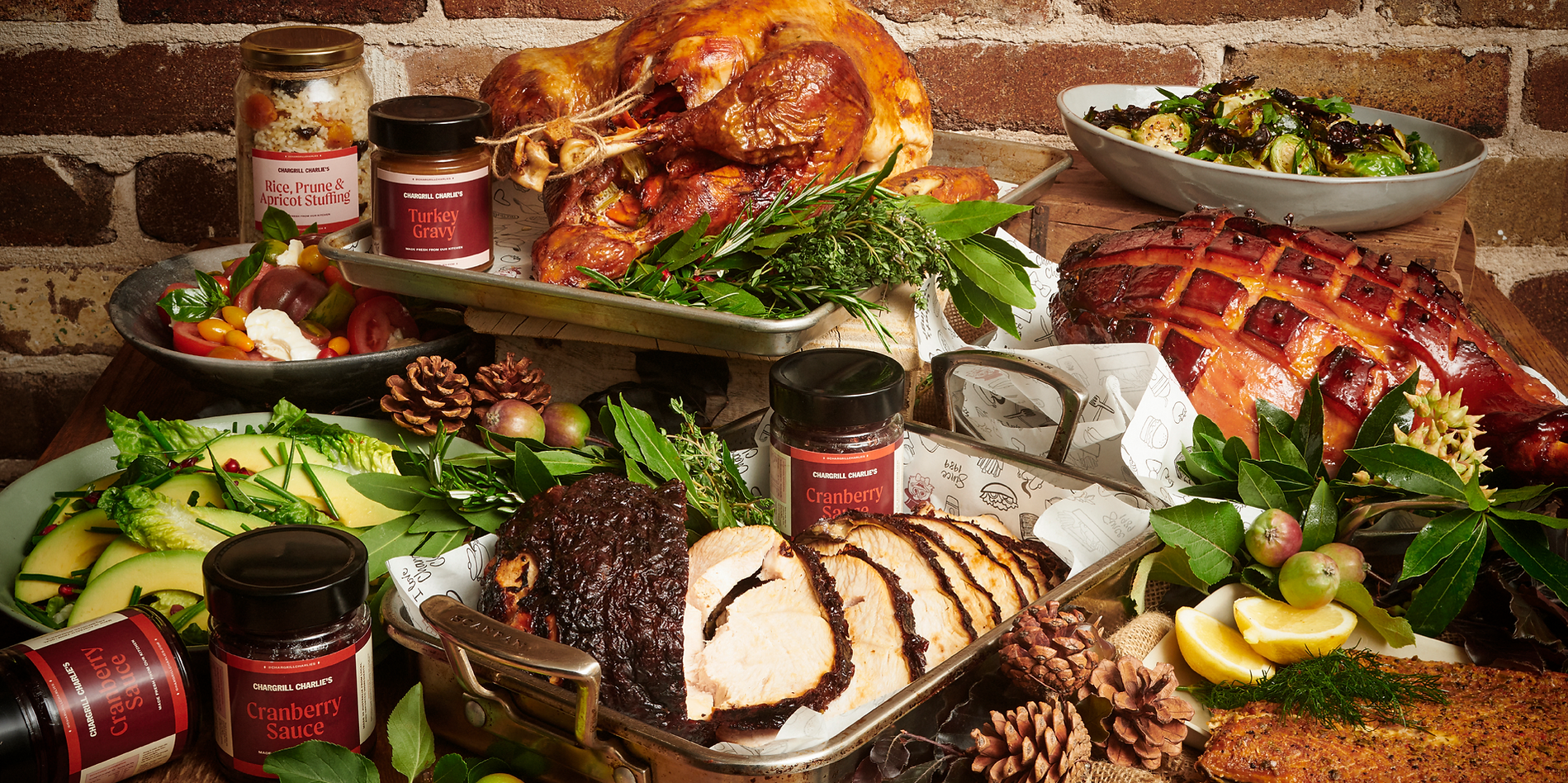 Overhead shot of a Thanksgiving feast with roasted turkey, ham, salads, cranberry sauce, and stuffing against a brick background.