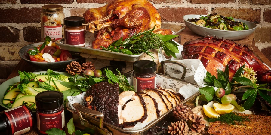 Overhead shot of a Thanksgiving feast with roasted turkey, ham, salads, cranberry sauce, and stuffing against a brick background.