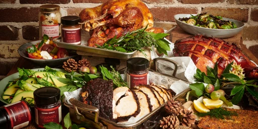 An abundant holiday feast featuring a roasted turkey, glazed ham, side dishes, and packaged condiments against a brick background.
