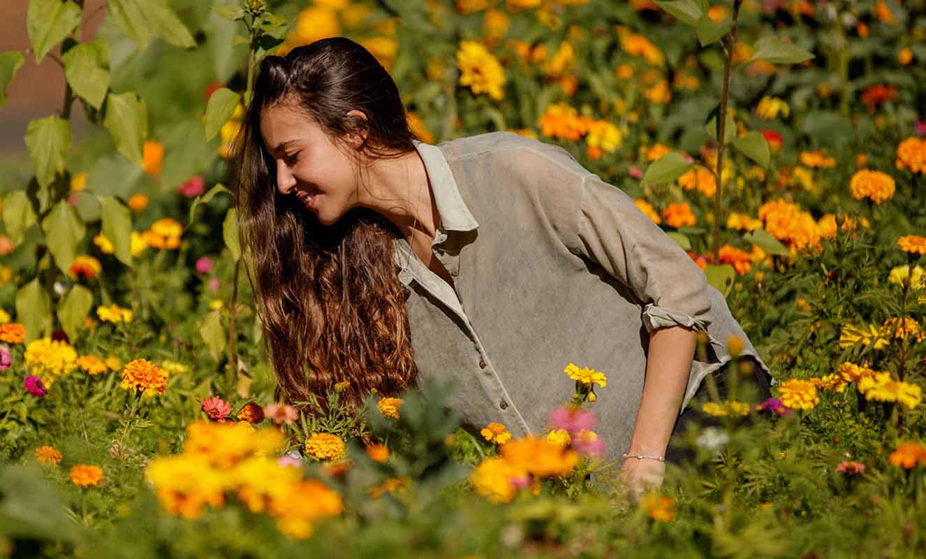 Young woman in a garden bending to look at yellow and orange marigold flowers on a sunny day with green foliage in the background.