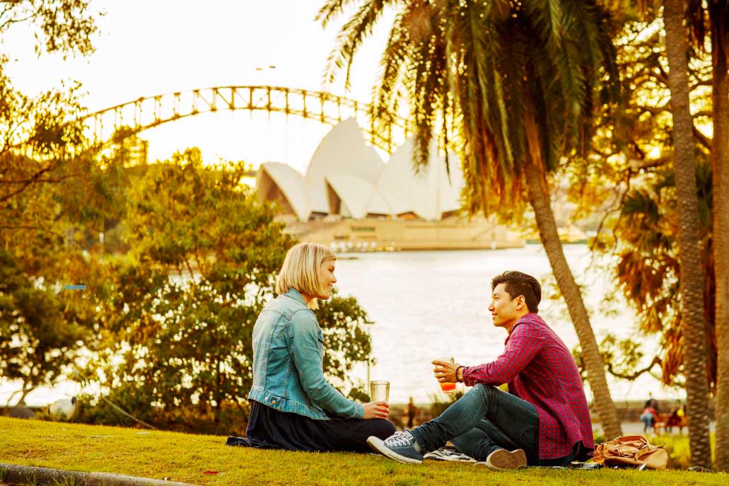 Couple sits on a grassy hill overlooking the Sydney Opera House and Harbour Bridge in golden light during daytime, creating a serene scene.