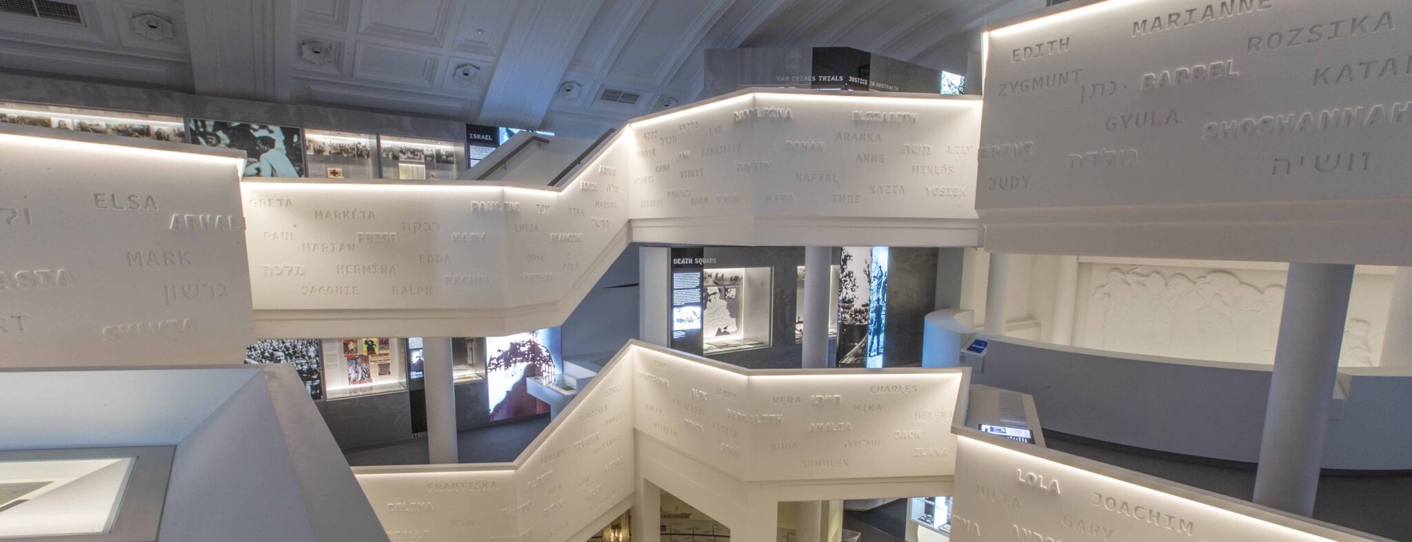 Interior view of a white museum exhibit with stairs, walls covered in names, informational displays, and interior architecture.