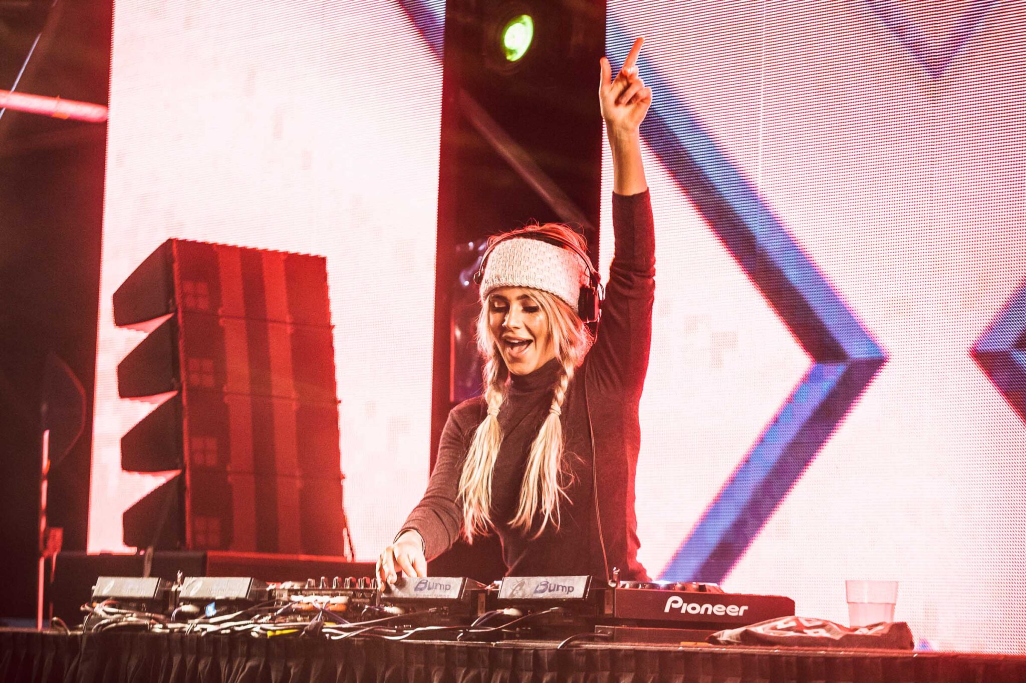 Blonde female DJ in a dark turtleneck and knitted headband raises her hand, working with Pioneer DJ equipment at an indoor music venue.