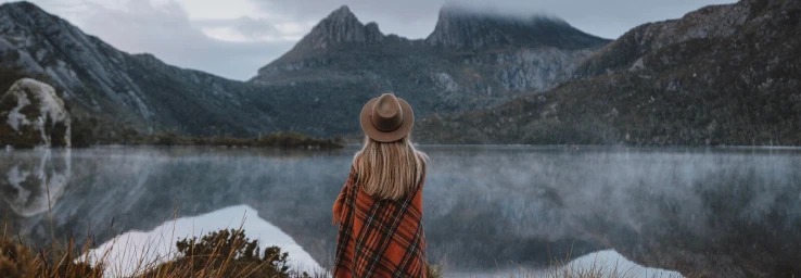 Woman wearing a brown fedora and a red patterned blanket poncho facing a misty lake and rugged mountain range