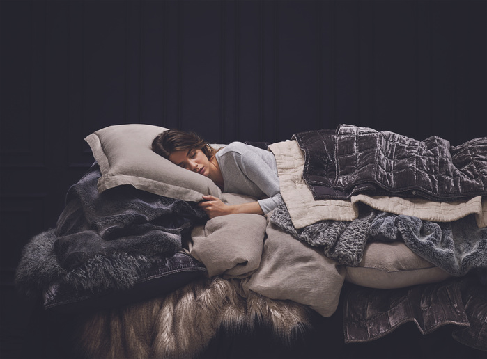 Adult woman sleeping on a bed covered in gray-toned pillows and blankets against a dark background. Indoor studio photograph.
