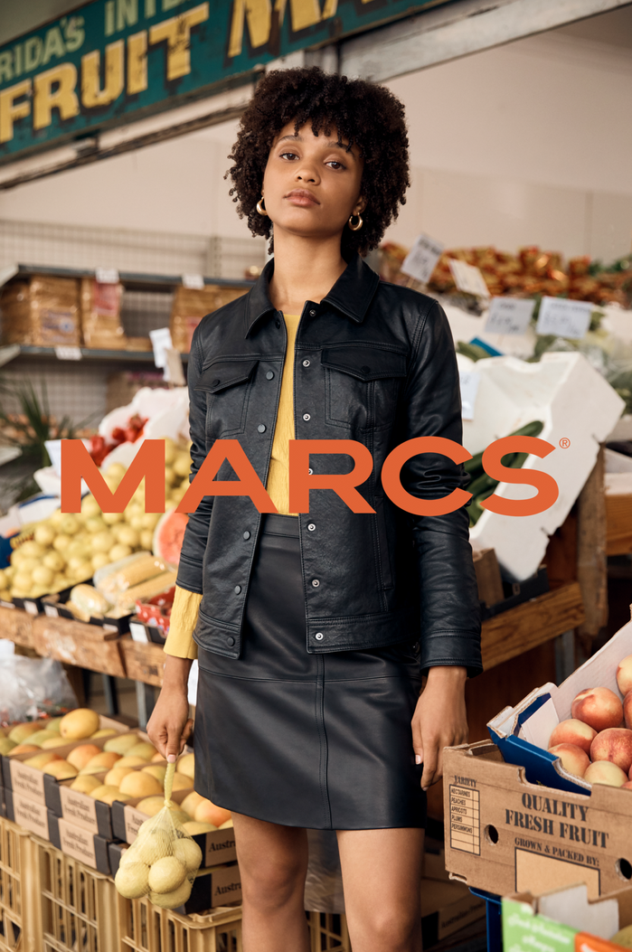 A brunette adult female poses in a black leather jacket and skirt with a background display of fruits and vegetables at a market.