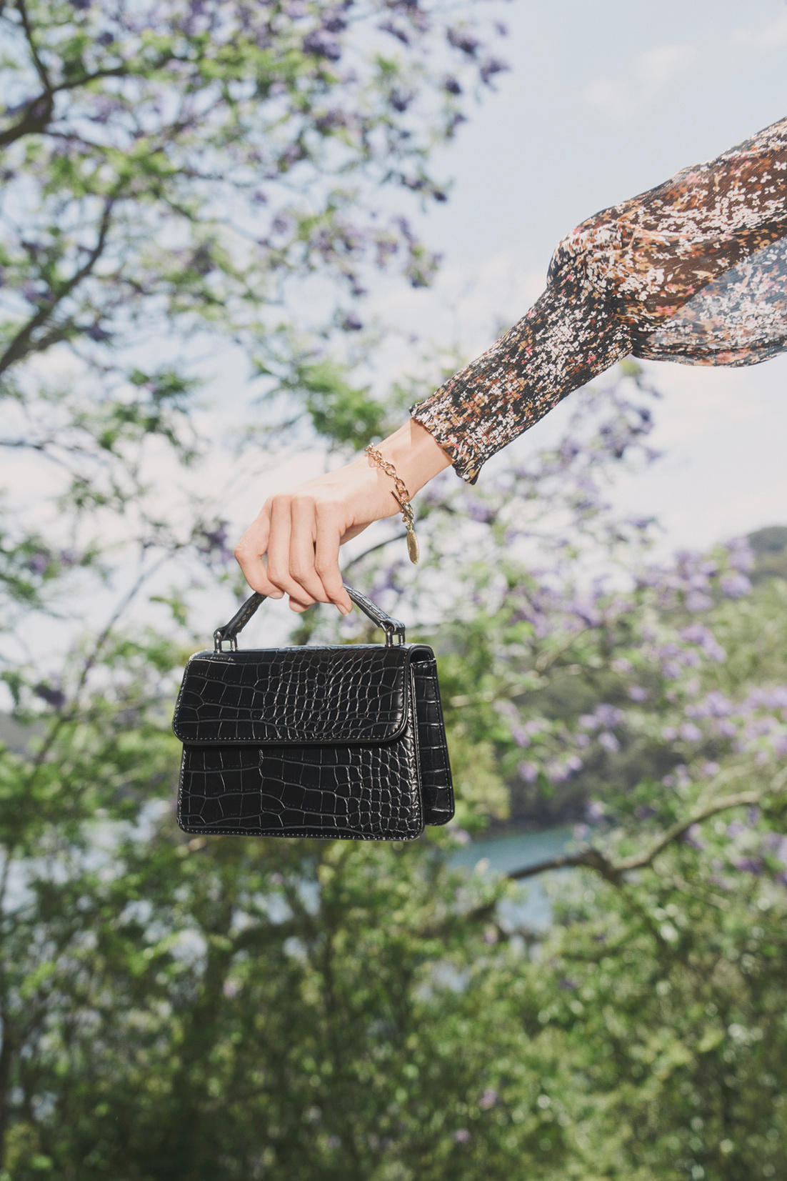 A black, textured leather top-handle bag is held up against a blurred outdoor background of green trees and purple jacaranda blossoms.