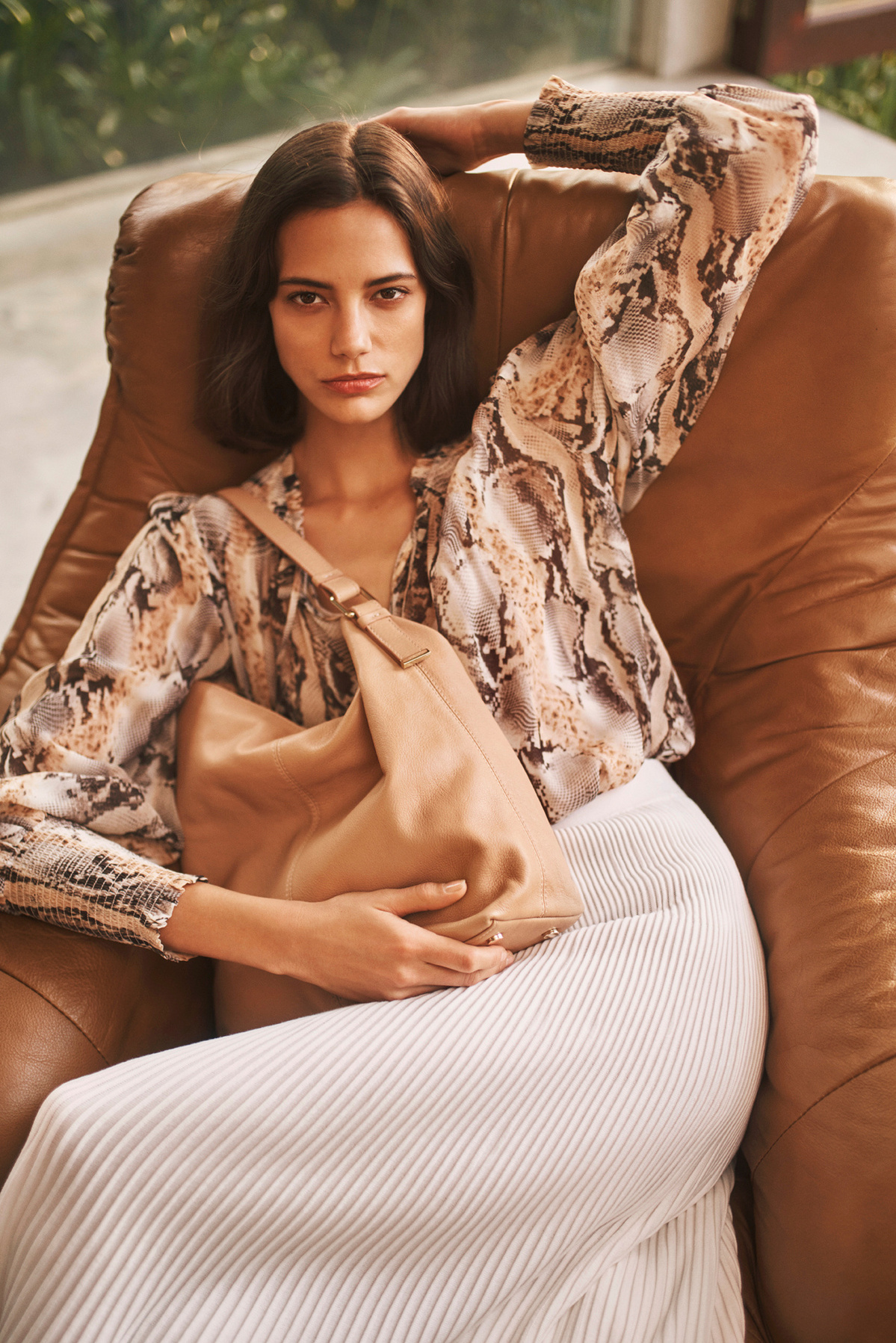 Indoor portrait of a woman with dark hair, wearing an animal print blouse and pleated skirt, seated in a brown leather chair and holding a tan handbag.